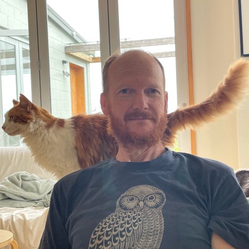 Adam in an owl t-shirt, with a ginger and white cat walking along the couch behind his shoulders